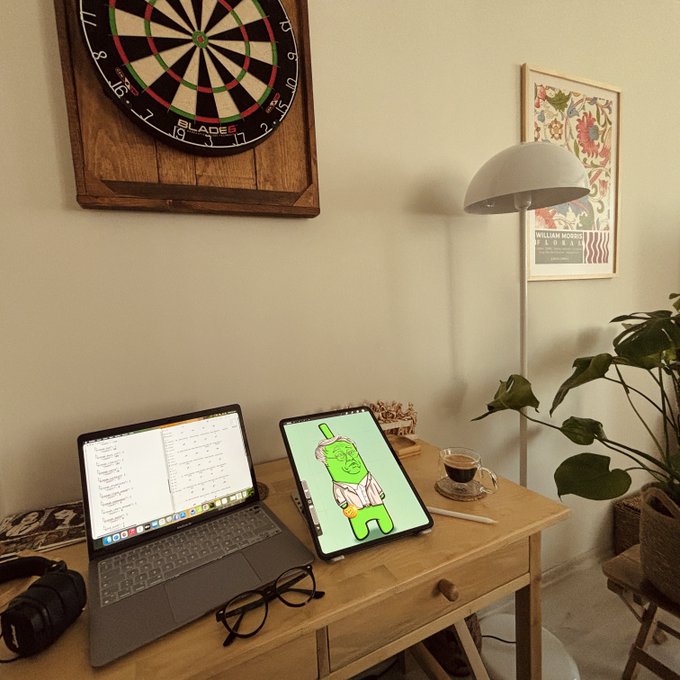 A cozy home office setup featuring a laptop and tablet on a wooden desk, with a dartboard on the wall above, a floor lamp, a potted plant, and a William Morris poster.