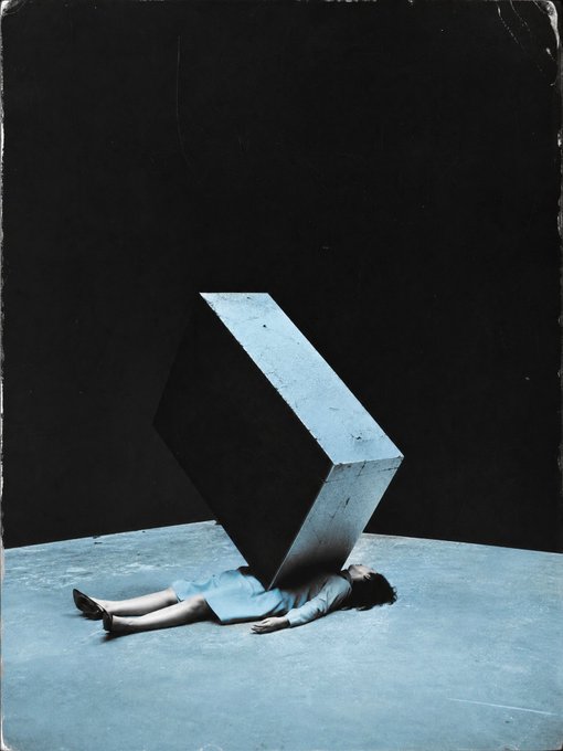 A surreal, dark artistic photograph showing a woman in a blue dress lying on a pale floor, her body seemingly crushed or supporting a massive, tilted concrete cube in a shadowy void.