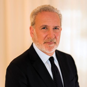 A professional headshot shows a gray-haired, bearded man in a dark suit and tie against a light background, consistent with a formal portrait of economist Peter Schiff, whose commentary on Kevin Warsh's continuation of inflationary monetary policy accompanies the image.