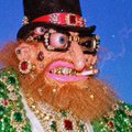 Low-resolution image showing a person with a large orange beard, sunglasses, red earrings, and a top hat, appearing to be a stylized or filtered portrait.