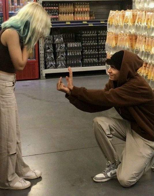A scene in a store showing a young man in a brown hoodie and beanie kneeling and making a finger gun gesture toward a person with blonde and teal hair who is looking down with their face covered.