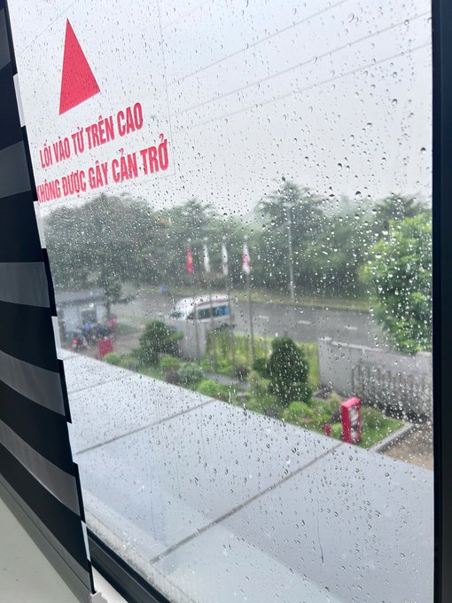 A rainy view through a window with water droplets, showing a warning sticker in Vietnamese ('LỖI VÀO TỪ TRÊN CAO / KHÔNG ĐƯỢC GÂY CẢN TRỞ') and a street scene with vehicles and greenery outside.