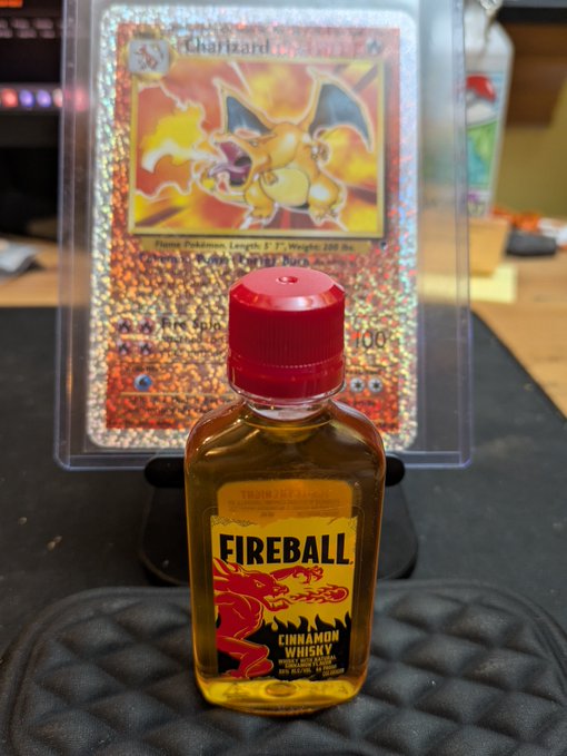 A miniature bottle of Fireball Cinnamon Whisky sitting in front of a holographic Charizard Pokémon trading card displayed in a protective case.