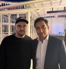 Two men pose together at what appears to be an indoor event venue, with one wearing a black cap and glasses in a dark sweater, and the other dressed in a dark blazer over a white shirt. The image relates to the post about an XRP Seoul event, capturing an unexpected meeting with Monica and Asheesh before the conference begins.