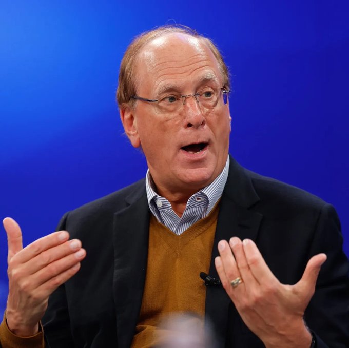 An older man with glasses, wearing a dark blazer over a brown sweater and striped shirt, speaks animatedly while gesturing with both hands against a blue backdrop—appearing to be Larry Fink, CEO of BlackRock. The image relates to the news of BlackRock clients purchasing $114.32 million worth of ETH, reflecting the firm's continued investment activity in Ethereum.