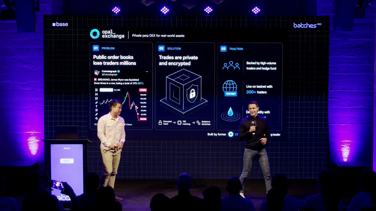 In this image, two men are standing on a stage with a large screen behind them displaying information about the @base accelerator program. The first man is wearing a white shirt, while the second man has his arms crossed. They appear to be presenting or discussing something related to the program. The background of the image features a black and purple color scheme, which complements the content displayed on the large screen behind them.