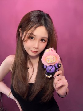 The image features a woman with long brown hair, wearing a black dress, holding up a pink stuffed animal. She appears to be posing for a photo or selfie, possibly sharing her excitement about the toy she's holding. The background of the image is a pink wall, which complements the color of the stuffed animal and adds a vibrant touch to the scene.