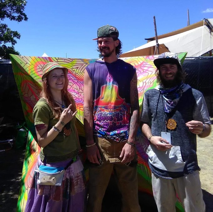 Three people pose at what appears to be a festival, standing in front of a colorful tie-dye backdrop with a tent visible in the background.