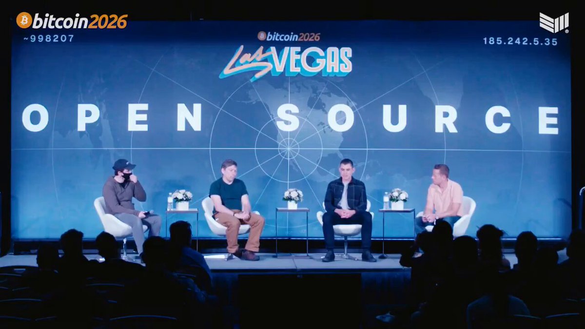 The image shows a group of four people sitting around a table with chairs arranged for them during an open source event. They are engaged in conversation about Bitcoin and its future, possibly discussing the implications of the signature scheme used by Bitcoin. The background features a large screen displaying the Open Source logo, indicating that this is likely an event related to Bitcoin or blockchain technology.