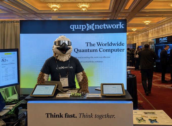 Conference booth photo showing someone in an eagle mascot costume wearing sunglasses at a 'Qup Network' display advertising 'The Worldwide Quantum Computer' with the slogan 'Think fast. Think together.'