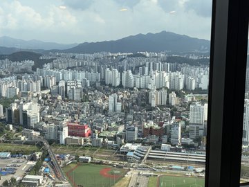 Aerial view from a high-rise window overlooking a dense cityscape with high-rise apartment buildings, lower urban sprawl, and mountains in the hazy background.