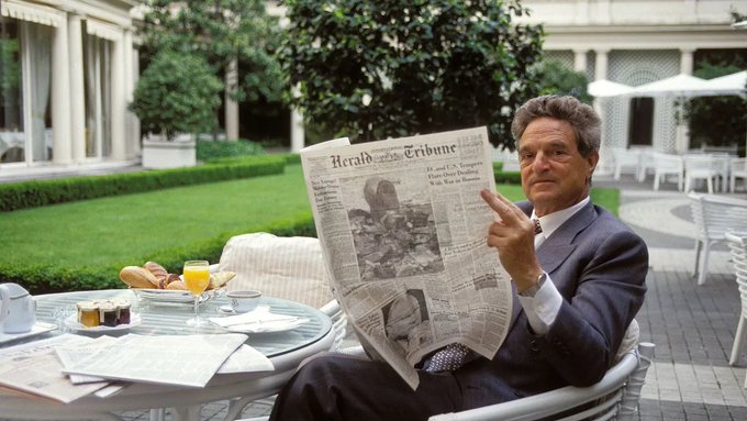 A man in a dark suit sitting at an outdoor patio table reading a copy of the 'Herald Tribune' newspaper, with a plate of pastries and orange juice in front of him.