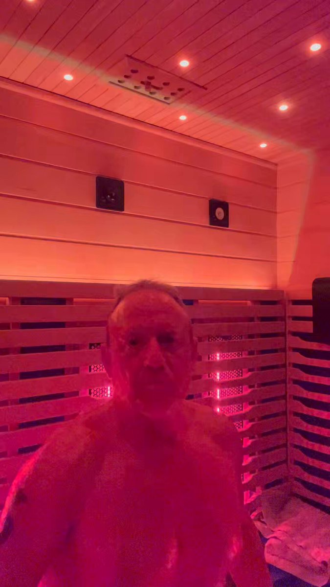 The image features an older man standing inside of a sauna, surrounded by wooden walls that are illuminated with red lights. The man appears to be posing for the camera while he enjoys his time in the sauna.