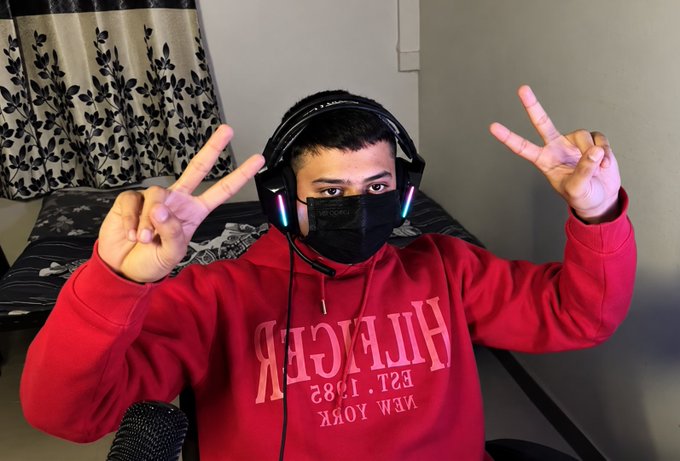 A person wearing a red Hilfiger hoodie, a black face mask, and a gaming headset poses with two peace signs in a bedroom.