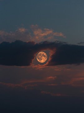 A luminous full moon glows through a break in dark, layered clouds, framed by wisps of cloud that resemble eyelashes, creating the appearance of a giant eye peering through the night sky. The image brings to life the evocative "moon eye" concept, with the surrounding clouds forming a natural eyelid against the deep twilight.