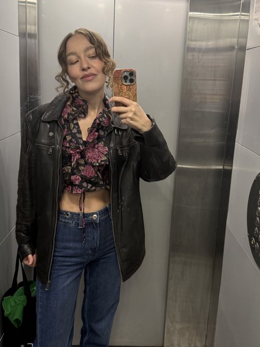 A woman takes a mirror selfie in a stainless-steel elevator, wearing a black leather jacket, floral top, and jeans while holding an orange-cased phone and a black-and-green bag. The photo serves as a casual personal check-in accompanying the upbeat post about waking up late, FOMO, Kimchi’s X giveaways, partying, and getting back to replying to friends.