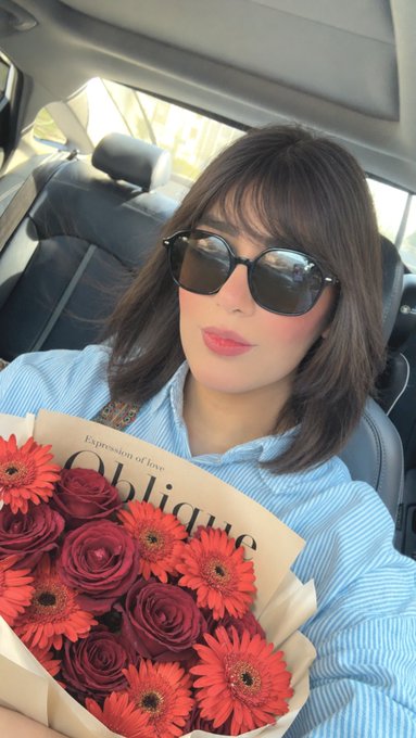 A woman wearing sunglasses and a blue striped shirt sits in a car holding a bouquet of red roses and orange gerberas wrapped in paper labeled 'Expression of love' and 'Oblique'.