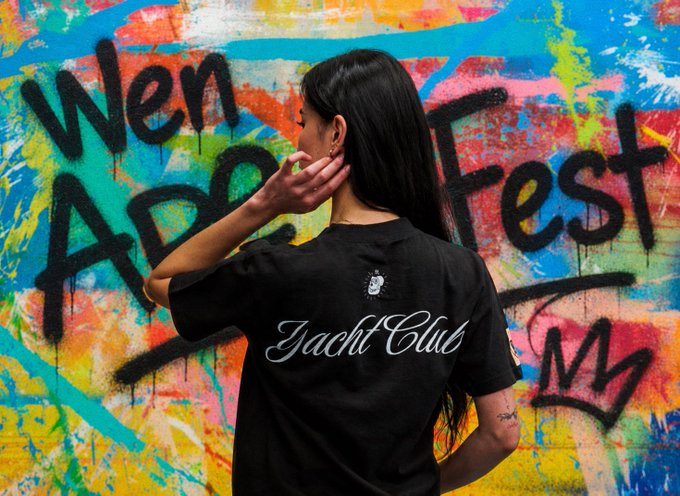 Woman with long black hair wearing a black 'Yacht Club' t-shirt, standing in front of a colorful graffiti wall with text reading 'Wen Abstract'.