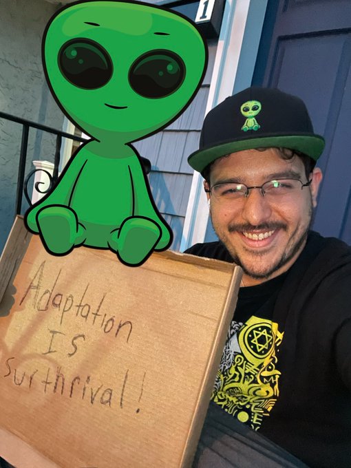 A selfie of a smiling man wearing glasses and a black cap with a green alien logo, holding a cardboard sign that reads "Adaptation Is Survival!" next to a digital green alien character.