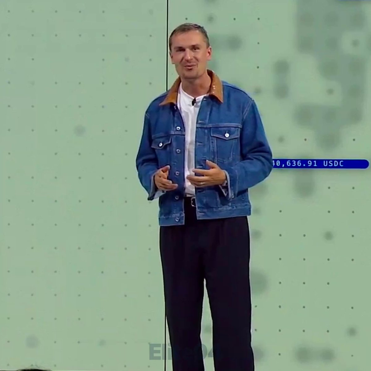 The image features a man wearing a blue denim jacket, black pants, and a white shirt standing against a green wall with a blue border. The man appears to be giving a speech or presentation, as indicated by his body language and the fact that he is looking directly at the camera.