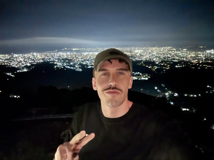 A nighttime selfie of a mustached man in a backward cap posing with a peace sign, overlooking a sprawling city skyline.