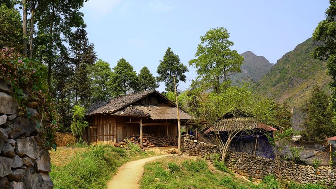 The image shows an old wooden house with a thatched roof situated at the bottom right corner of the frame, surrounded by trees and mountains. The photo has been edited to include a caption above the house, which reads "Good morning everyone Woke up. Checked @sleepagotchi #Sleepagotchi #SleepToEarn #Bestpartofthemorning #Everymorning #Frens".