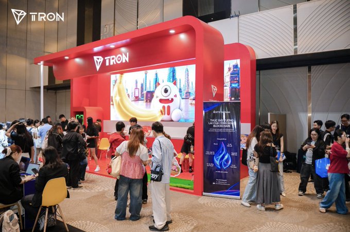 A TRON blockchain exhibition booth at what appears to be a crypto conference, featuring a large red display with the TRON logo, digital screens, and crowds of attendees exploring the space.