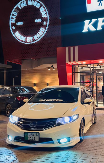 A white Honda sedan parked at night outside a KFC restaurant, with a circular neon sign reading 'MAO THE MADDEST MADE MY HAND' and a 'SINISTER' windshield banner.