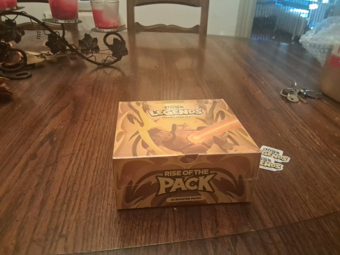 A Pokémon TCG 'Rise of the Pack' booster box sits on a wooden table next to keys, candles, and a decorative centerpiece.