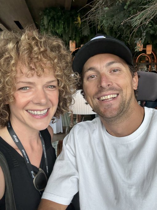 A smiling selfie of a couple, with a woman with curly blonde hair wearing a black top and lanyard next to a man in a black cap and white t-shirt, at what appears to be an outdoor restaurant or event venue.