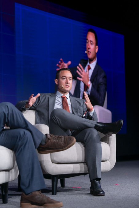 The image shows a man sitting in a chair, giving a speech or presentation while wearing a suit and tie. The setting appears to be an event where he is speaking about tokenized securities, DeFi, and other financial topics. In the background, there are two men standing behind him, possibly listening to his speech or waiting for their turn to speak.