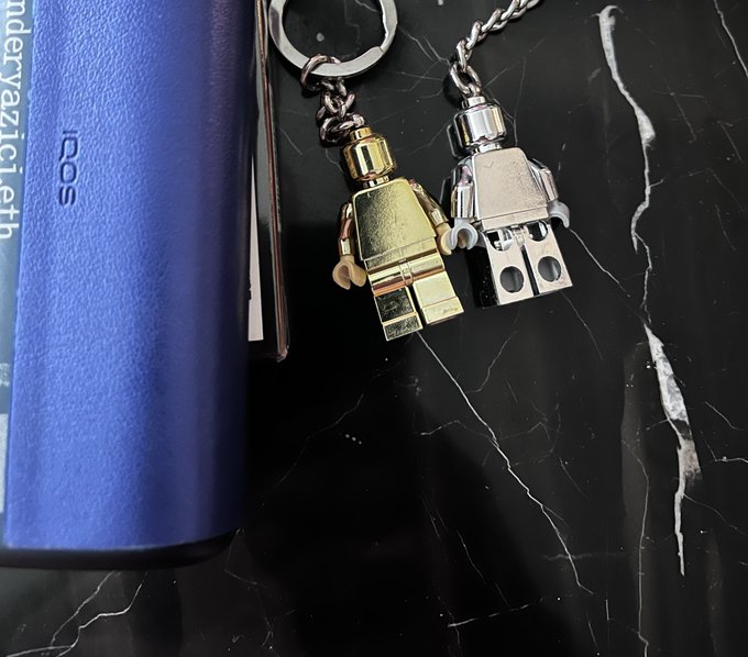 A photo of two Lego minifigure keychains (one gold, one silver) hanging next to a blue IQOS device on a black marble surface, with text 'anderyazici.eth' visible.