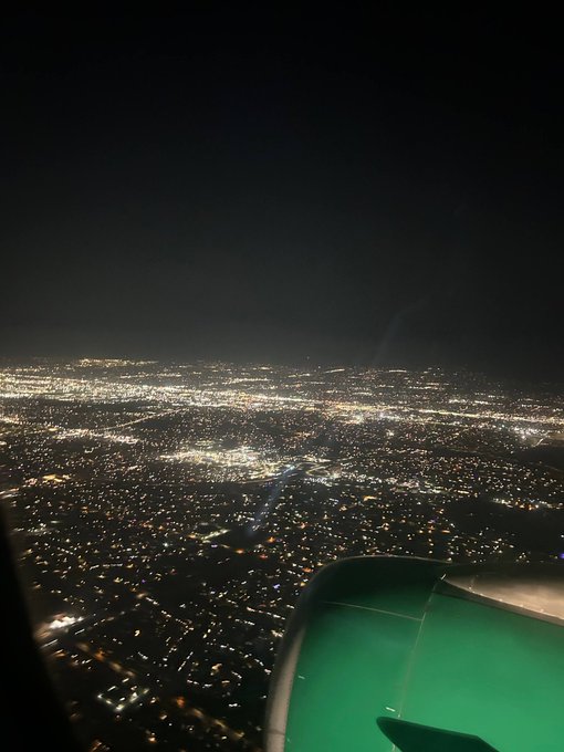 An aerial nighttime view of a sprawling city from an airplane, with the green-lit engine visible in the foreground.