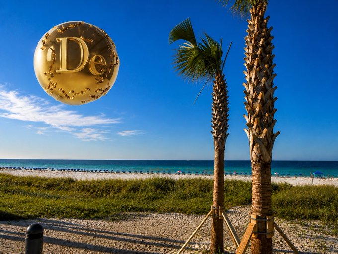 A golden Dogecoin coin with the "D" logo floats over a sunny beach scene with palm trees and the ocean.