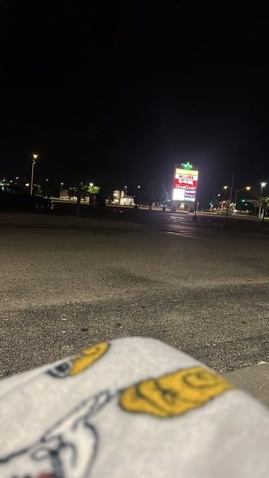 A nighttime view of a parking lot with an illuminated sign in the distance, with a patterned white towel or blanket visible in the foreground.