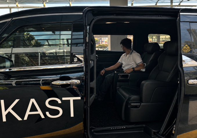A man sits in the back of a black Alphard luxury van with the word 'KAST' visible on the side, the sliding door open.
