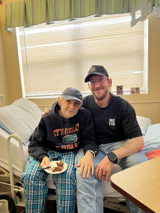 An elderly man wearing a black 'IT'S GREAT' hoodie and blue plaid pants sits on a hospital bed eating cake, while a younger man in a black 'Fresh Ink' cap and t-shirt sits beside him with his arm around the older man.