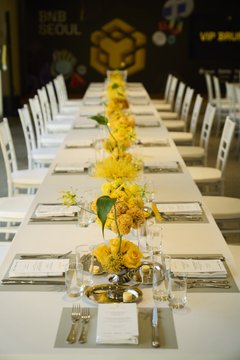 A long banquet table at the BNB Seoul VIP Brunch is set with white linens, place settings, wine glasses, and a striking centerpiece of tall yellow and green floral arrangements. The event signage visible in the background reinforces the upscale "BNB Seoul VIP Brunch" gathering atmosphere.