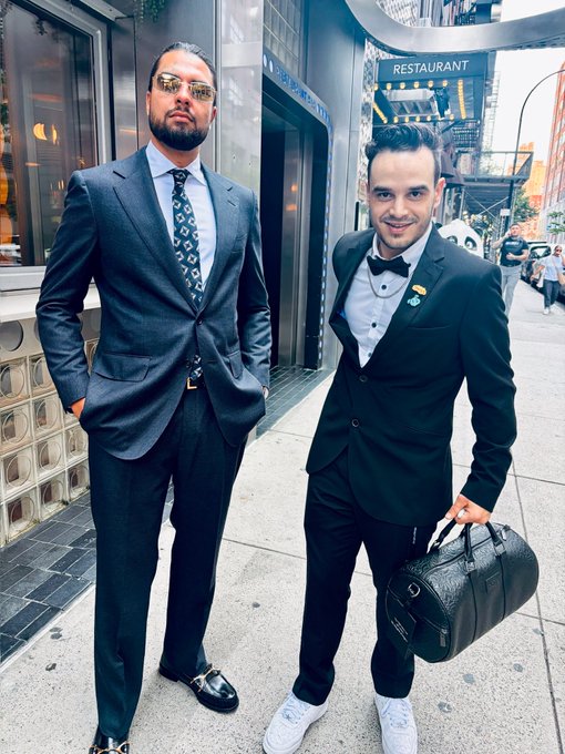 Two well-dressed men in suits stand on a city sidewalk outside a restaurant; one wears a blue suit and sunglasses, while the other wears a black tuxedo with a bow tie and holds a black duffel bag.