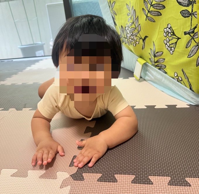 A baby with a blurred face lies on a play mat doing a tummy time exercise, with a yellow floral curtain in the background. The image illustrates the post's mention of spending the day at home playing with "ベビ吉" (Baby-kichi), the user's affectionate nickname for their baby, after going to vote on an election day.