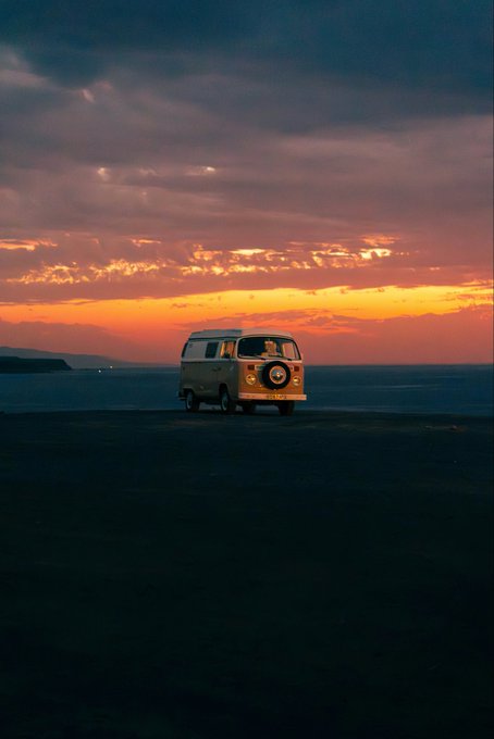 A vintage Volkswagen camper van parked on a sandy beach at sunset, with dramatic pink and orange clouds illuminating the sky above the dark ocean horizon.