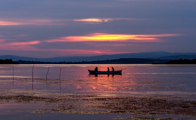 A scenic photograph of two people in a small boat silhouetted against a colorful sunset over a calm lake, with rolling hills in the background.