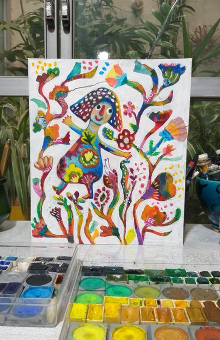 A colorful, whimsical painting of a figure among flowers and plants sits on a desk in front of an open watercolor palette with various paint colors visible in its compartments.