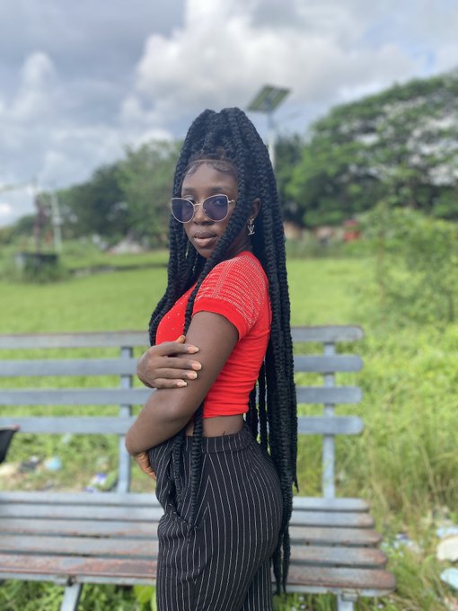 A young woman with long black braided hair, sunglasses, a red top, and pinstriped pants poses with crossed arms in front of a park bench.