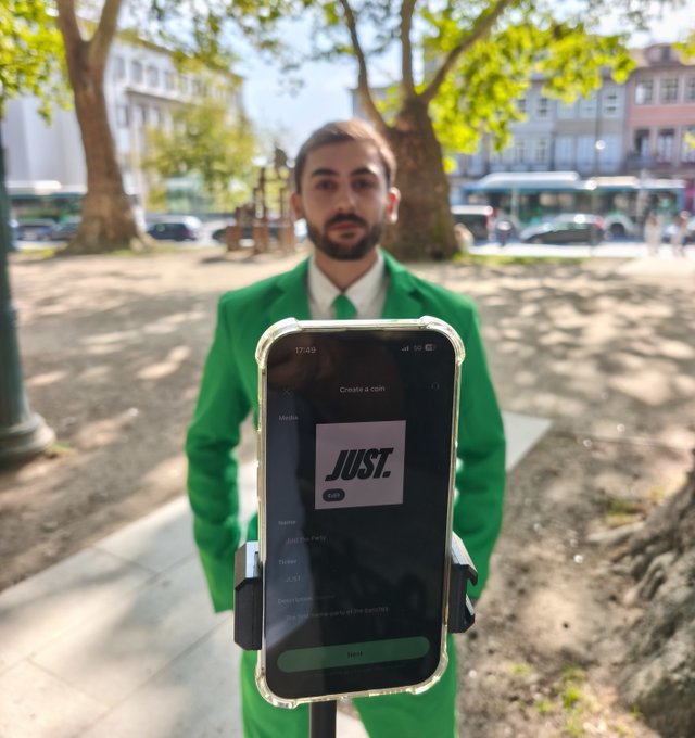 A man in a green suit is promoting the "$JUST" coin and its "meme-party of the trenches" by showing his phone screen, which displays "JUST." as the coin logo and "JUST" as the ticker.