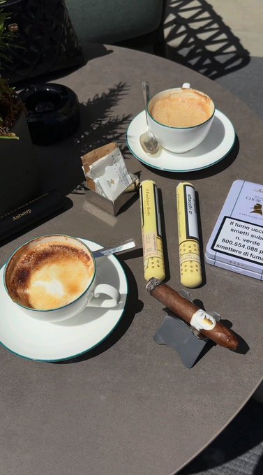 Two cups of coffee on saucers sit on an outdoor table alongside a pair of yellow binoculars, an ashtray, a business card, and a cigar resting on a stand.