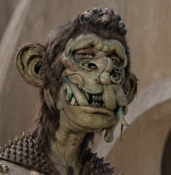 A close-up of a creature with greenish, textured skin, pointed ears, prominent tusks, and a spiked collar.
