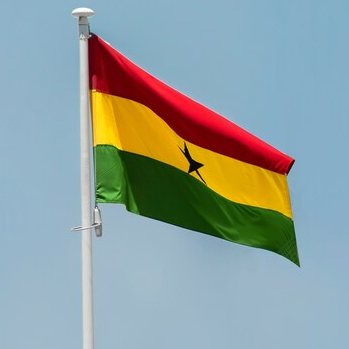 A photograph of the Ghanaian flag (red, yellow, and green horizontal stripes with a black star in the center) waving on a flagpole against a clear sky.