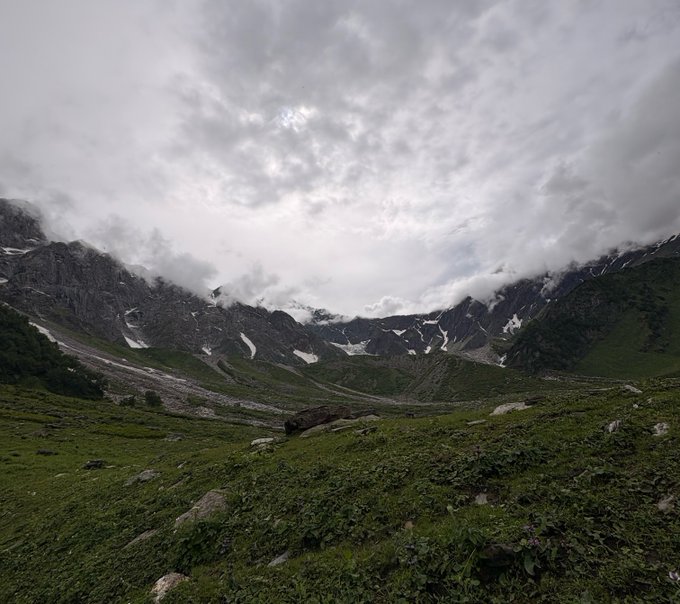A scenic landscape of a mountain valley with green grassy foreground, rocky peaks with patches of snow, and an overcast sky filled with gray clouds.