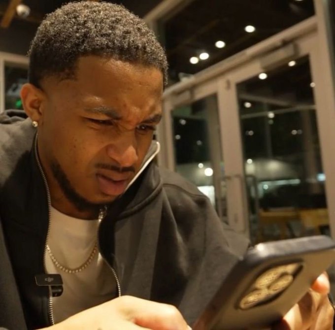 A young man with a short haircut, wearing a black jacket and gold chain, squints with a confused or frustrated expression while looking at his smartphone with a triple-camera lens setup.